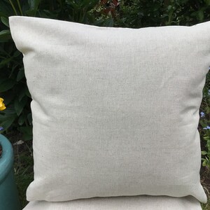 May include: A square, light beige pillow with a textured surface. The pillow is made of a woven fabric and has a simple, classic design. The pillow is outdoors, with greenery and flowers in the background.