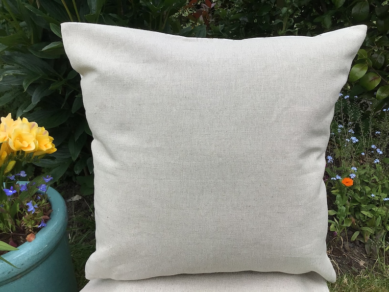 May include: A square, light beige pillow with a textured surface. The pillow is made of a woven fabric and has a simple, classic design. The pillow is outdoors, with greenery and flowers in the background.
