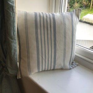 May include: Square pillow with a cream background and vertical blue stripes. The pillow is resting on a white windowsill next to a light green curtain. The pillow is a decorative home accent.