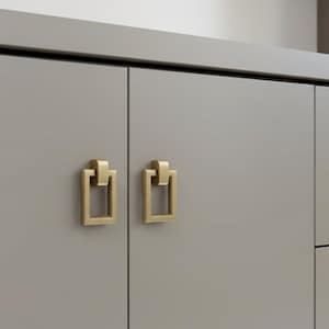 May include: Close-up of two gold-colored cabinet pulls with a rectangular design. The pulls are mounted on a gray cabinet door. The hardware has a modern, minimalist aesthetic, suitable for kitchen or bathroom cabinetry.