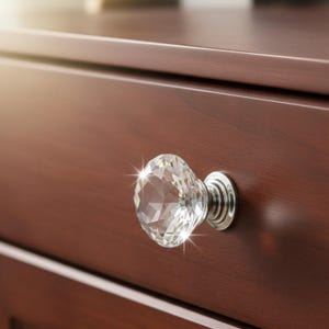 May include: Close-up of a crystal cabinet knob with a faceted design, mounted on a dark brown wooden drawer. The knob has a silver-colored base and reflects light, creating a sparkling effect. The wood grain is visible.