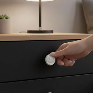 May include: A white, rose-shaped drawer pull on a black drawer. The drawer is part of a nightstand with a wooden top. A hand is touching the drawer pull. A lamp and a small potted plant are visible in the background.