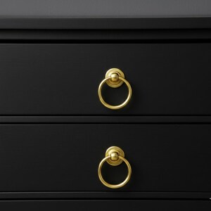 May include: Close-up of a black dresser with two drawers. Each drawer features a gold-colored ring pull. The pulls are circular with a decorative backplate. The dresser has a simple, modern design.