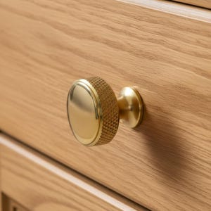 May include: Close-up of a gold-colored cabinet knob with a textured, rounded edge and a smooth, oval center. The knob is attached to a light-colored wooden cabinet, showcasing the natural wood grain.
