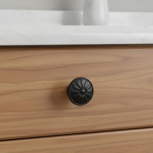 May include: A black, round cabinet knob with a floral design is mounted on a light brown wooden drawer. The knob has a detailed, textured surface and a central circular element. The background includes a white countertop and a white vase.