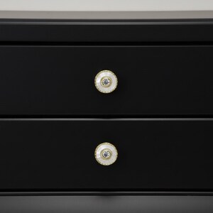 May include: Close-up of a black dresser with two drawers. Each drawer has a decorative knob with a white center, a gold-colored rim, and a blue-gray center detail. The knobs add a touch of elegance to the dark furniture.