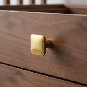 May include: Close-up of a wooden drawer with a square, gold-colored knob. The knob is centered on the drawer, and the wood grain is visible. The drawer is part of a larger piece of furniture.