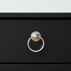 May include: Close-up of a black drawer with a silver ring pull. The ring pull is circular and attached to a circular backplate. The drawer is part of a larger piece of furniture.