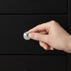 May include: A shiny, silver-colored cabinet knob held in front of a black cabinet. The knob is round with a slightly domed top and a flat base. The cabinet has a simple, modern design.