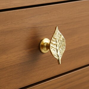 May include: A gold-colored leaf-shaped drawer pull with intricate detailing. The pull is mounted on a wooden drawer, showcasing the hardware's design and finish. The leaf design adds a touch of nature-inspired elegance to the furniture.