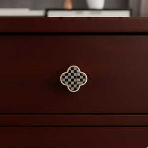 May include: A close-up of a dark brown wooden cabinet with a four-leaf clover-shaped drawer pull. The drawer pull has a gold-colored trim and a black and white checkered pattern in the center. The cabinet has a smooth, polished finish.