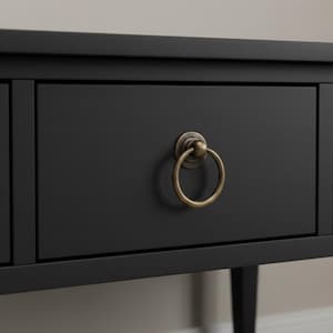 May include: Close-up of a black wooden drawer with a brushed metal ring pull. The drawer is part of a larger piece of furniture, with visible legs and a top. The ring pull is a warm brass color, contrasting with the dark drawer.