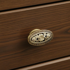 May include: An antique brass oval drawer pull with a floral rose design. The drawer pull is mounted on a dark brown wooden surface. The decorative hardware features a detailed, vintage aesthetic.