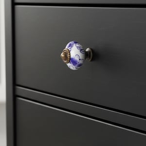 May include: A close-up of a black drawer with a decorative knob. The knob is white with blue floral patterns and a brass-colored base. The drawer is part of a larger black cabinet.