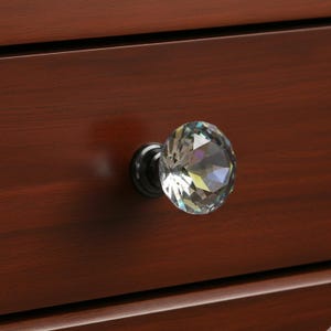 May include: A faceted crystal knob with a black metal base is attached to a dark brown wooden drawer. The crystal knob reflects light, creating a sparkling effect. The wood grain is visible.