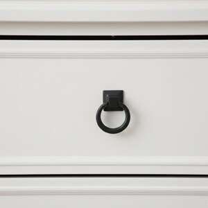 May include: Close-up of a white drawer with a black ring pull. The ring pull is square at the base and circular in the center. The drawer has a recessed panel design.