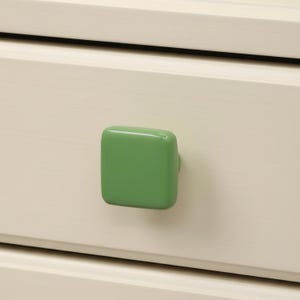 May include: Close-up of a cream-colored drawer with a square, green knob. The knob is centered on the drawer front, providing a pop of color against the neutral background. The drawer appears to be part of a larger piece of furniture.