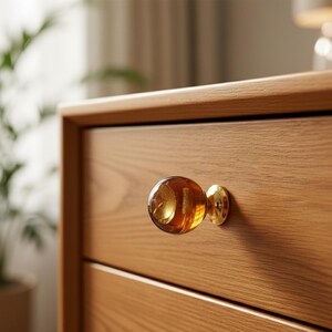 May include: Close-up of a wooden drawer with a decorative amber glass knob and a gold-colored metal base. The drawer is made of light brown wood. The knob has a round shape and a textured interior.
