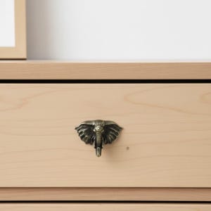 May include: A bronze-colored elephant head drawer pull mounted on a light wood drawer. The elephant head has detailed features, including large ears and a trunk. The drawer is part of a light-colored wooden dresser or cabinet.