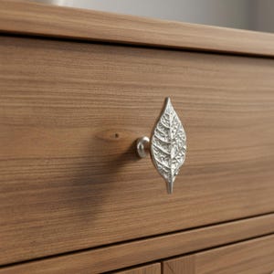 May include: A silver-colored leaf-shaped drawer pull is mounted on a wooden cabinet. The drawer pull is detailed with leaf veins and has a small, round base. The cabinet has a natural wood grain finish.