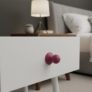 May include: A white nightstand with a pair of round, magenta-colored drawer pulls. The nightstand has a minimalist design with a clean, smooth surface and wooden legs. A lamp and a bed are visible in the background.