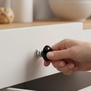 May include: A black faceted cabinet knob with a chrome base is attached to a white drawer. The knob is being pulled open. The background includes a wooden countertop and a white cabinet.