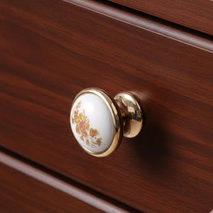 May include: A close-up of a drawer knob with a white ceramic face featuring a floral design in yellow and pink. The knob has a gold-colored metal rim and base, set against a dark brown wooden drawer.
