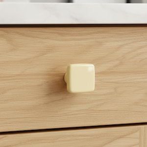 May include: A square, pale yellow cabinet knob is mounted on a light-colored wooden drawer. The knob has rounded edges and a smooth, glossy finish. The wood grain of the drawer is visible.