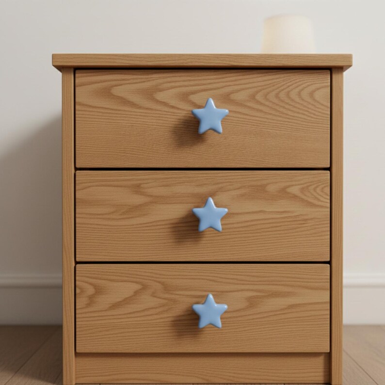 Puede incluir: Una mesita de noche de madera de tres cajones con tiradores en forma de estrella azul claro. La madera clara tiene una veta visible. Un peque&ntilde;o objeto cil&iacute;ndrico blanco est&aacute; encima.