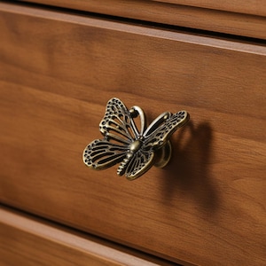 May include: A bronze-colored butterfly-shaped cabinet knob is mounted on a wooden drawer. The knob has intricate details, including openwork wings and a detailed body. The wood grain is visible, and the overall aesthetic is vintage.