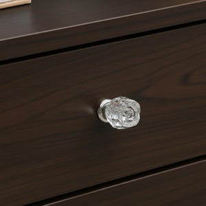 May include: Close-up of a dark brown wooden drawer with a clear, faceted glass knob. The knob has a floral design and a silver-colored base. The wood grain is visible on the drawer front.