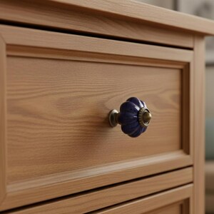 May include: Close-up of a wooden dresser drawer with a blue and gold floral knob. The drawer is made of light brown wood with visible grain. The knob is a dark blue color with gold accents and a silver base.