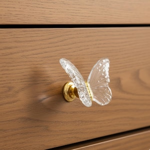 May include: A clear glass butterfly cabinet knob with gold-colored hardware. The butterfly design is detailed and delicate, with a gold accent on the body. The knob is mounted on a wooden cabinet.