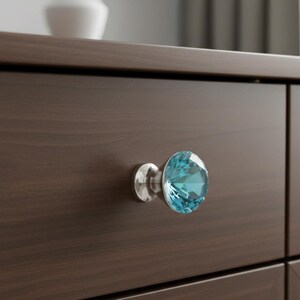 May include: A close-up of a dark brown wooden drawer with a single, decorative knob. The knob features a faceted, light blue crystal set on a silver-colored base. The drawer's wood grain is visible.