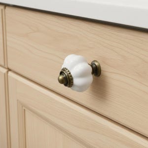 May include: A white, ceramic cabinet knob with a fluted design and antique brass hardware. The knob is mounted on a light wood cabinet door. The hardware includes a decorative ring and a small, round base.