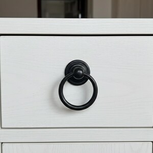 May include: Close-up of a white wooden drawer with a black ring pull. The drawer has a light wood grain texture. The ring pull is circular and attached to a round base.