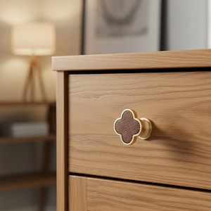 May include: A close-up of a wooden drawer with a decorative clover-shaped knob. The knob has a brown leather center and a gold-colored metal frame. The drawer is made of light-colored wood.