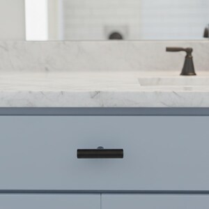 May include: A close-up of a light blue bathroom cabinet with a dark, cylindrical drawer pull. The countertop is white with gray veining, and a dark faucet is visible in the background. The cabinet is part of a bathroom vanity.