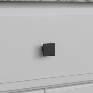May include: A square, matte black cabinet knob is mounted on a light gray cabinet door. The knob is centered on the door, providing a modern, minimalist aesthetic. The cabinet door has a recessed panel design.