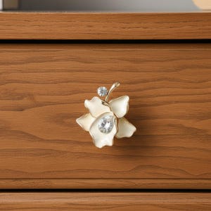 May include: A decorative flower-shaped cabinet knob with a cream-colored enamel finish and gold accents. The knob features a large, clear crystal center and a smaller crystal on top. The knob is mounted on a wooden drawer.