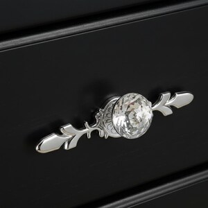 May include: Close-up of a black drawer with a crystal knob and silver decorative accents. The knob is round and faceted, reflecting light. The silver accents have a leaf-like design.