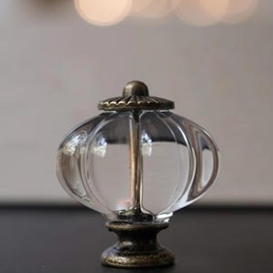 May include: Clear glass cabinet knob with a ribbed, pumpkin-shaped design. The knob has a bronze-colored base and top, and a central metal rod. The glass is transparent, allowing light to pass through. A decorative hardware element.