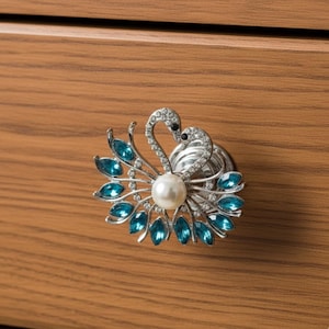May include: A decorative cabinet knob featuring two swans with silver-colored bodies and blue gemstone accents. The knob has a pearl in the center and is mounted on a wooden surface.