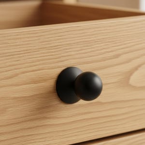 May include: Close-up of a wooden drawer with a black, round knob. The knob is attached to the drawer front with a circular base. The wood grain is visible, and the drawer appears to be part of a larger piece of furniture.