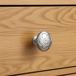 May include: Close-up of a silver metal drawer knob with a floral design. The knob is attached to a light brown wooden drawer. The knob has a rounded shape with a flower-like pattern in the center.