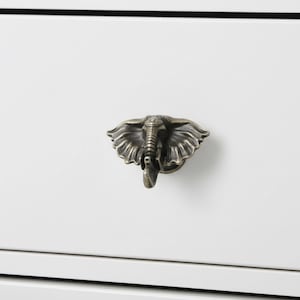 May include: A bronze-colored elephant head drawer pull mounted on a white cabinet. The elephant head is detailed with textured skin and large ears. The cabinet has a clean, modern design.