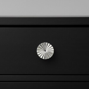 May include: A silver, starburst-shaped drawer pull is mounted on a black drawer. The drawer pull has a radial design with multiple ridges emanating from the center. The black drawer has a horizontal line at the bottom.