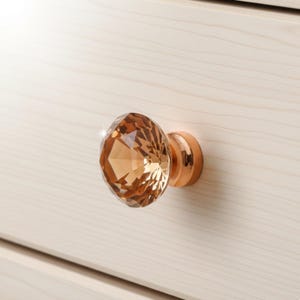 May include: A faceted, amber-colored crystal cabinet knob with a rose gold base. The knob is mounted on a light-colored wooden drawer, showcasing its decorative design and reflective qualities. The hardware adds a touch of elegance.