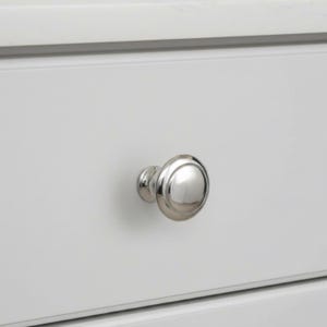 May include: Close-up of a polished, silver-colored cabinet knob. The knob is round with a slightly domed face and a short, cylindrical base. It is mounted on a white cabinet door, with a clean, minimalist aesthetic.