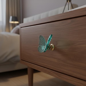 May include: A teal butterfly-shaped drawer pull with a brass base is mounted on a brown wooden nightstand. The butterfly has detailed wing patterns. The nightstand has a mid-century modern design with tapered legs.